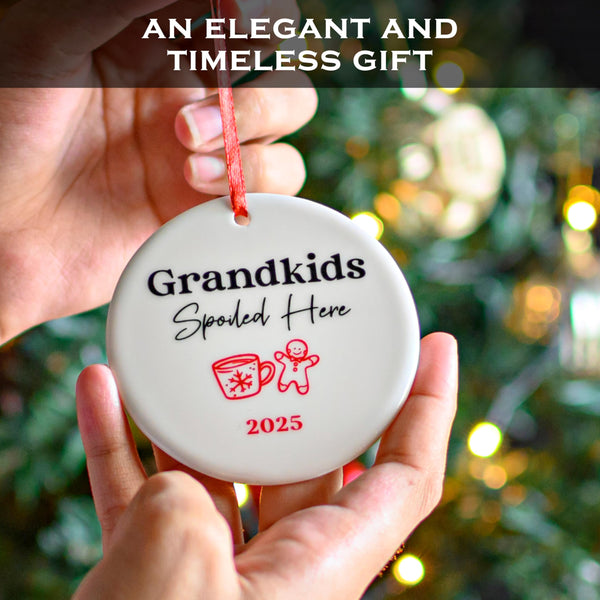 Round ornament with 'Grandkids Spoiled Here 2025' text held in front of a blurred Christmas tree.