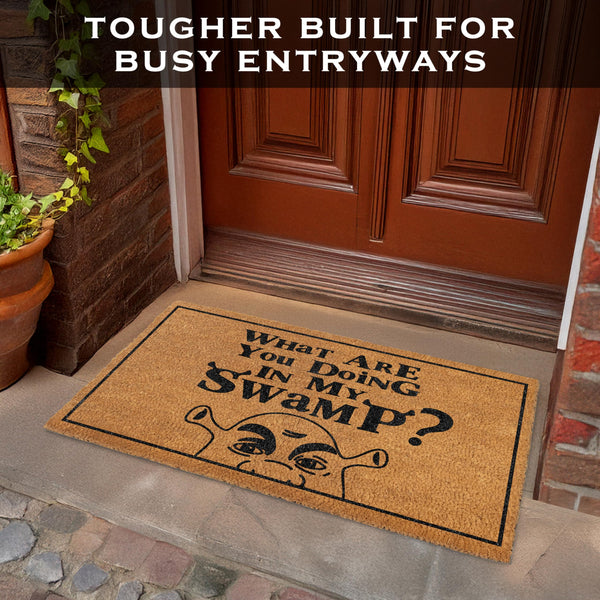 Doormat with humorous text on a front door step, featuring a plant and wooden door.